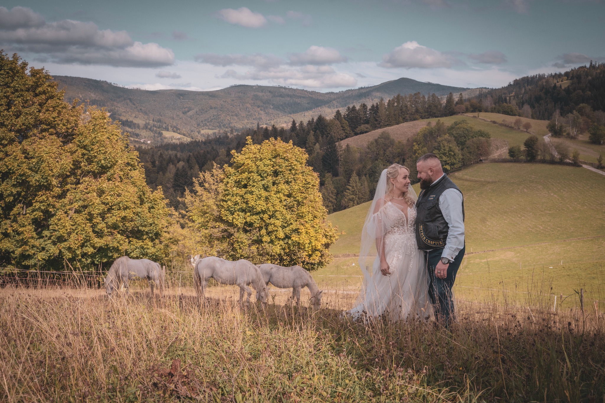 Para młoda w plenerze z końmi na tle malowniczych gór, panna młoda w koronkowej sukni, pan młody w kamizelce skórzanej.