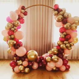 Okrągła, złota konstrukcja udekorowana girlandą z balonów w odcieniach różu, czerwieni i złota, ustawiona na drewnianej podłodze z kremową zasłoną w tle, tworząca dekorację eventową.
