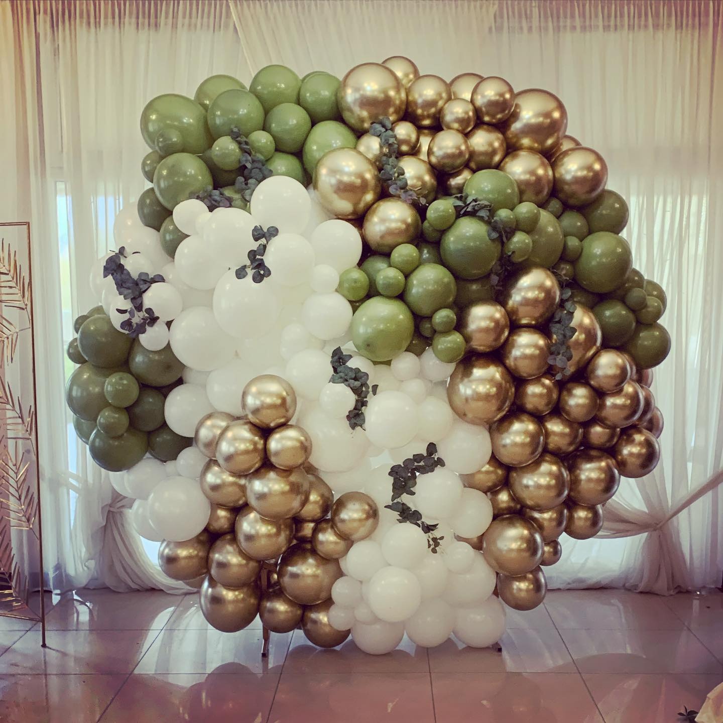 Dekoracja z balonów w kolorach złotym, białym i oliwkowym, ułożona w kształt kuli z dodatkiem gałązek eukaliptusa, na tle jasnych zasłon.