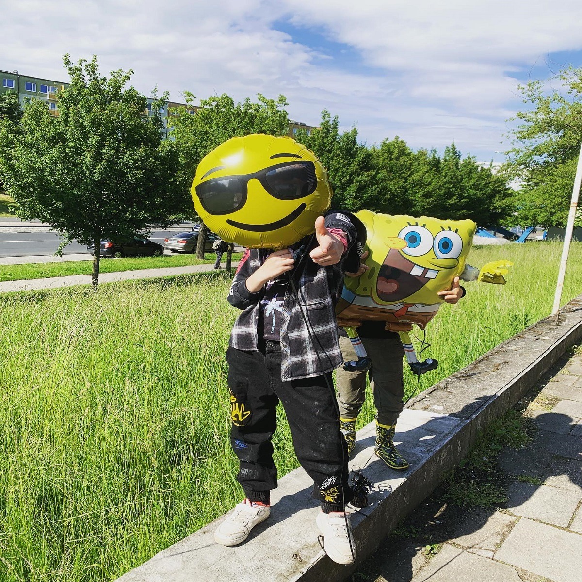 Dziecko z żółtym balonem emoji w okularach przeciwsłonecznych, pokazujące kciuk w górę, obok drugiego dziecka trzymającego balon Spongeboba na tle zielonej trawy i bloku mieszkalnego.