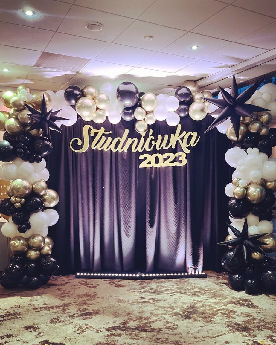 Dekoracja studniówki 2023 z czarnym tłem, złotym napisem, oraz girlandą z czarnych, złotych i białych balonów z gwiazdami po bokach.