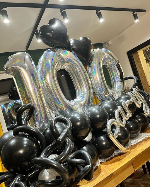 Dekoracja balonowa na 100. urodziny z czarnymi sercami, srebrnymi cyframi i napisem 'Party' na drewnianym stole z koronkową serwetą.