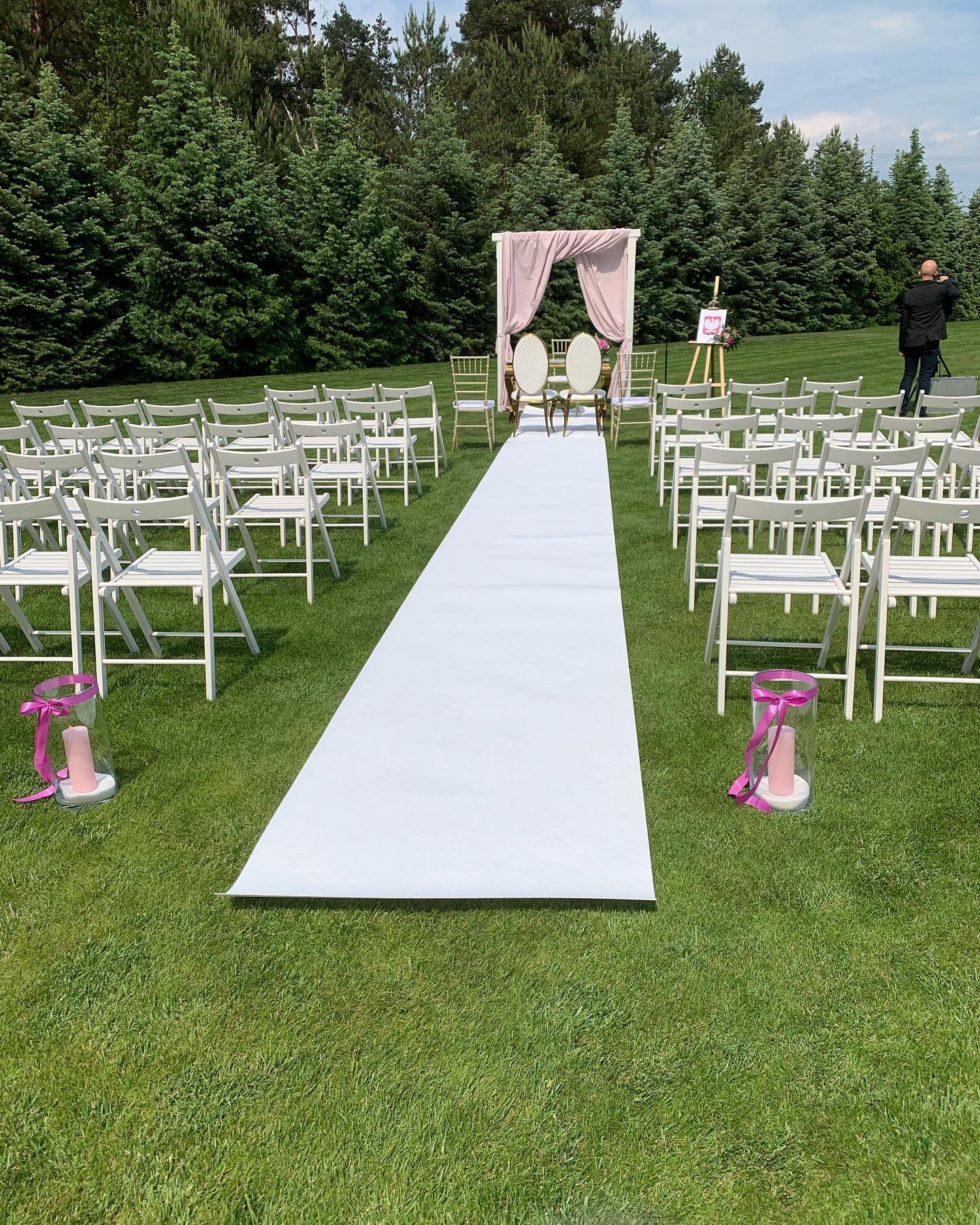 Przygotowane miejsce na ślub w plenerze: biały dywan, krzesła dla gości, ozdobny łuk z różową tkaniną, złote krzesła dla pary młodej, dekoracje ze świecami i różowymi wstążkami, fotograf...