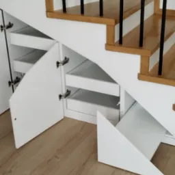 Biała zabudowa szafkowa pod schodami z otwartymi drzwiczkami i szufladami, widoczne drewniane schody z czarnymi balustradami.