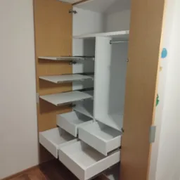 Otwarta szafa wnękowa z jasnego drewna z białymi półkami i szufladami na metalowych prowadnicach, widoczne zawiasy drzwiowe i drewniana podłoga.