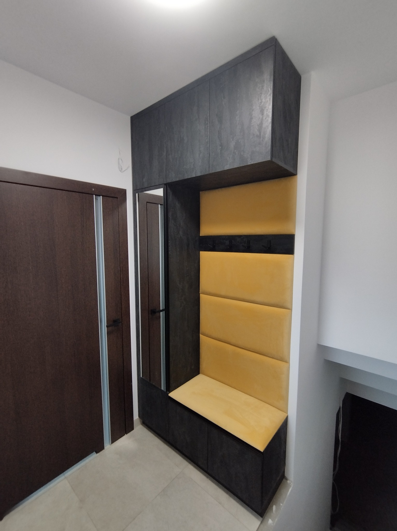 Wnęka w przedpokoju z szafkami, lustrem i siedziskiem z panelami w kolorze miodowym. Szafki i obudowa w kolorze grafitowym z widoczną fakturą betonu.