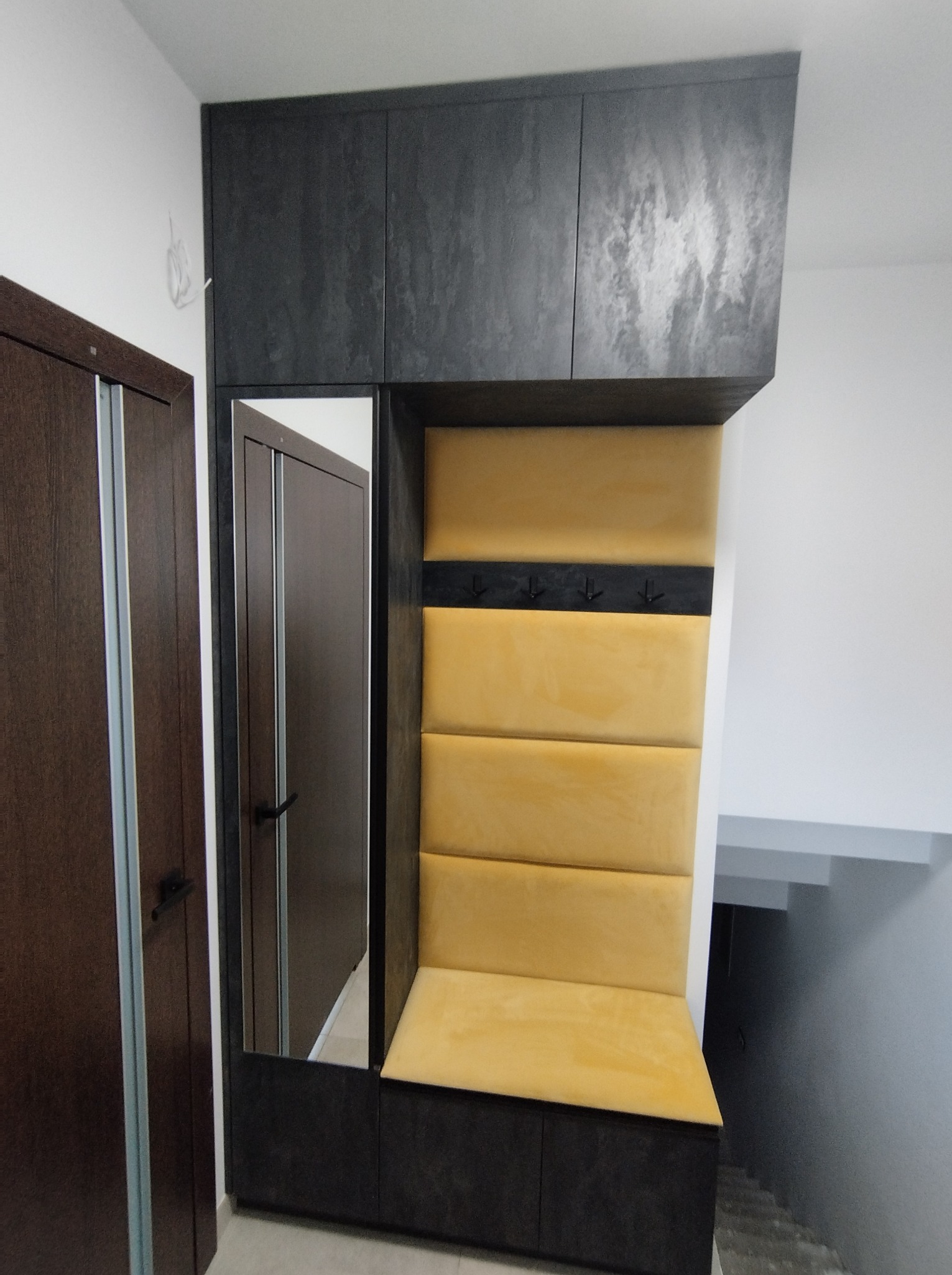 Wnęka wejściowa z szafkami, lustrem i tapicerowanym siedziskiem w odcieniach szarości i żółci, widok na klatkę schodową.