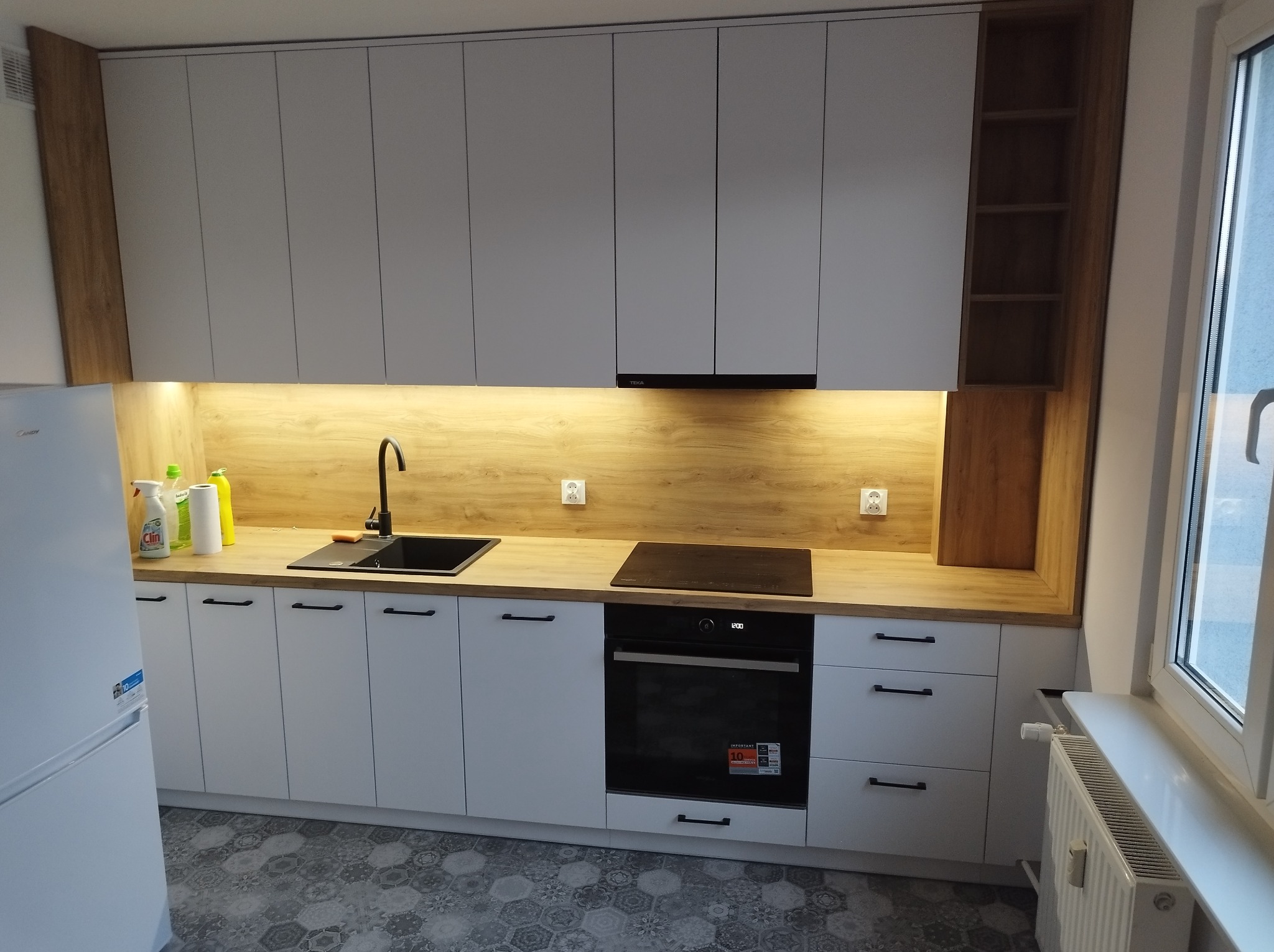 Jasna kuchnia w Myślachowicach z białymi frontami szafek i drewnopodobnym blatem, czarny zlew i płyta indukcyjna, widoczny fragment białej lodówki i grzejnik pod oknem.