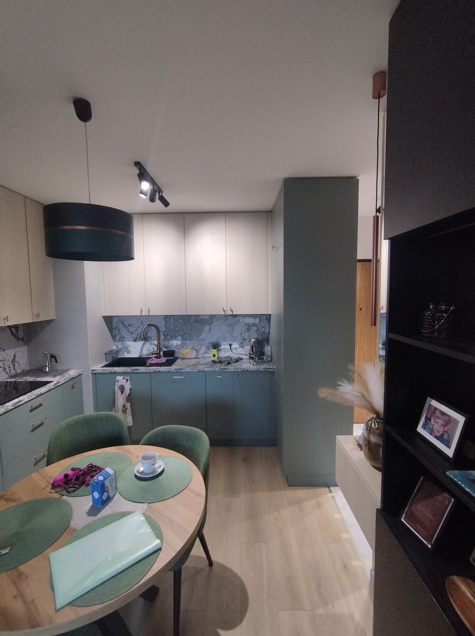 Nowoczesna kuchnia w odcieniach zieleni i beżu z marmurowym blatem, okrągły stół z zielonymi podkładkami i filiżanką, widok z perspektywy wnętrza.