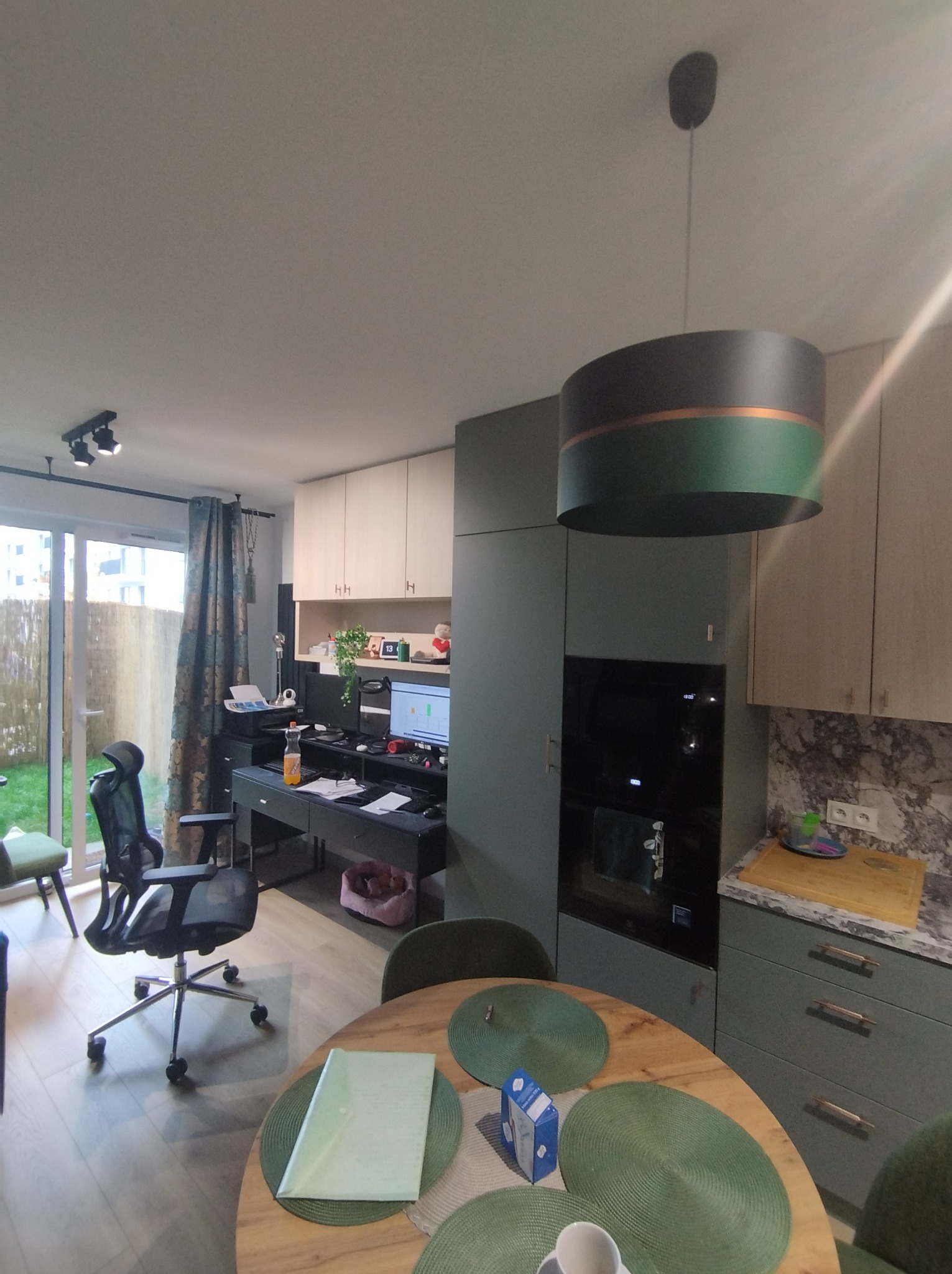 Wnętrze mieszkania z aneksem kuchennym w odcieniach zieleni i beżu, z okrągłym stołem z podkładkami i widokiem na biurko z komputerem i wyjściem na ogród.
