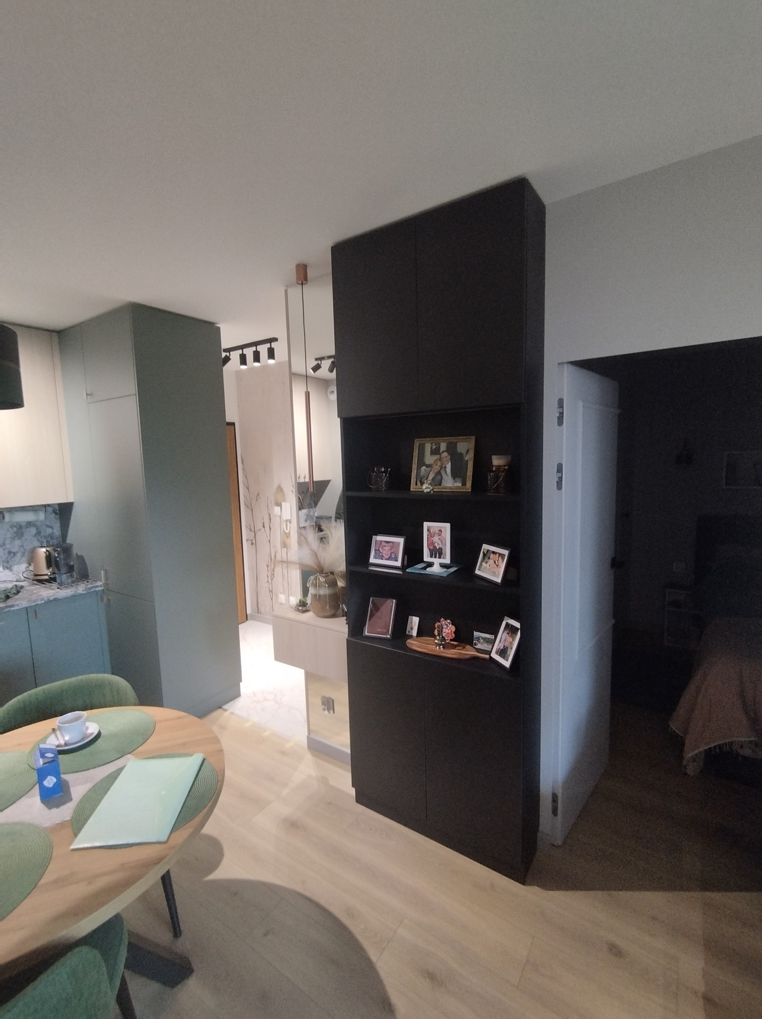 Ciemna szafa w zabudowie z otwartymi półkami na zdjęcia i dekoracje, oddzielająca kuchnię od sypialni, widok na stół z zielonymi podkładkami i filiżanką.