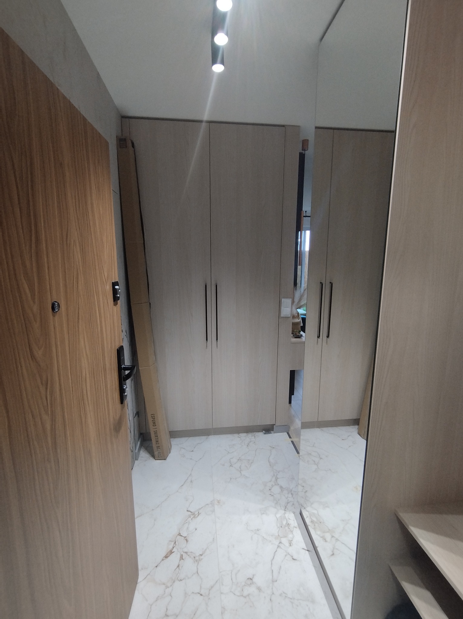Jasna szafa wnękowa z długimi, czarnymi uchwytami, widoczna obok otwartych drzwi wejściowych i kartonu z listwami przypodłogowymi, na marmurowej podłodze.