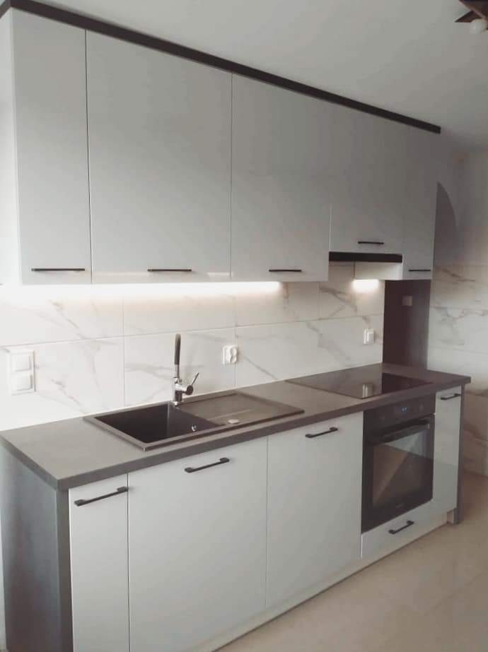 Nowoczesna, jasna kuchnia z białymi szafkami, ciemnym blatem i czarnymi uchwytami, marmurowe płytki na ścianie, oświetlenie LED pod szafkami górnymi, czarny zlew i płyta indukcyjna.