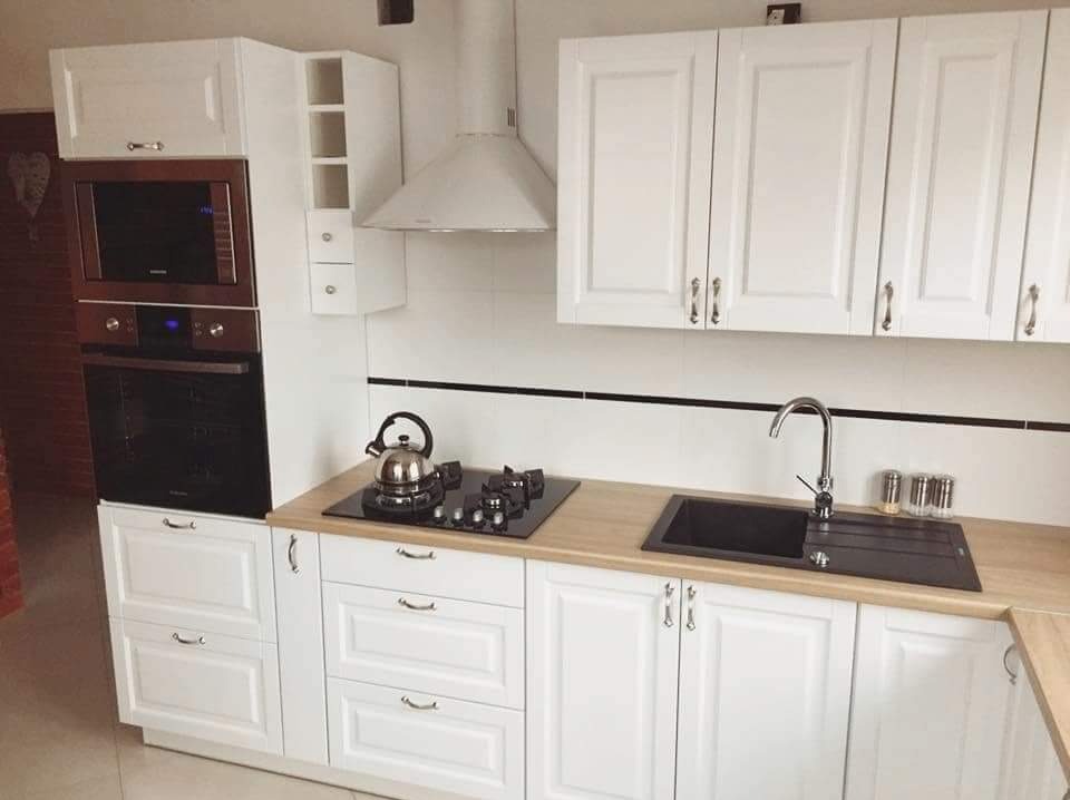 Biała kuchnia z drewnianym blatem, zabudowanym piekarnikiem i mikrofalówką, czarnym zlewem i płytą gazową z czajnikiem.