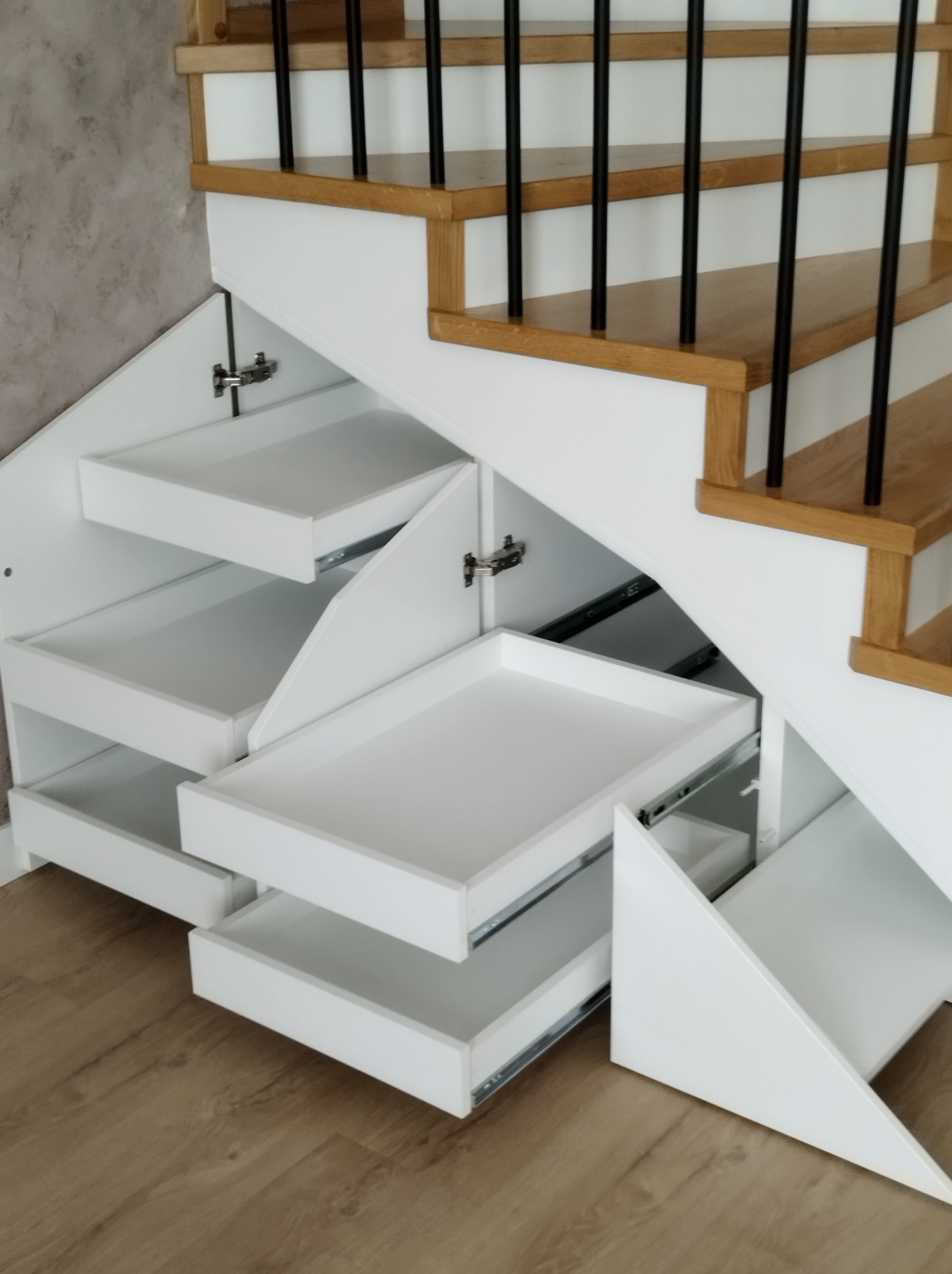 Białe, wysuwane szuflady pod schodami z drewnianymi stopniami i czarnymi balustradami, widoczne okucia meblowe.