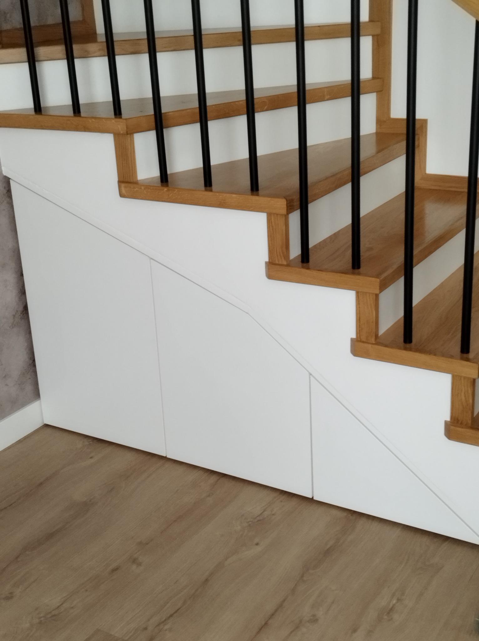 Schody drewniane z czarnymi balustradami i zabudową wnęki pod schodami z białymi, gładkimi frontami.