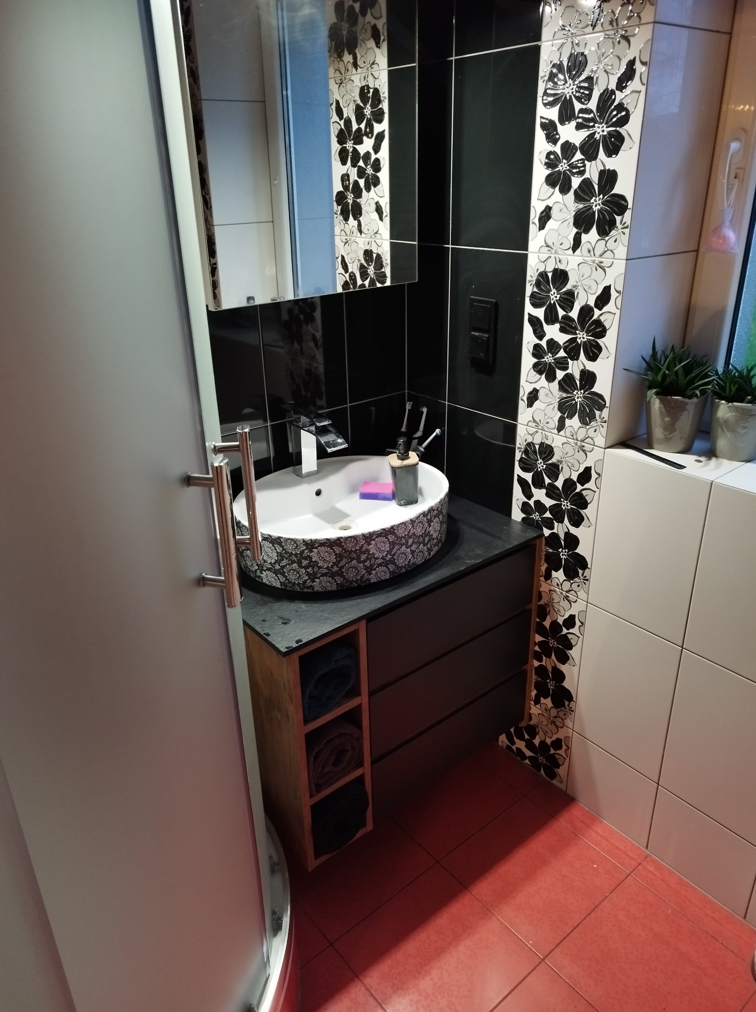 Nowoczesna łazienka z okrągłą umywalką w kwiatowy wzór, czarnymi płytkami z motywem kwiatowym, czerwoną podłogą i szafką z drewnianymi półkami na ręczniki.