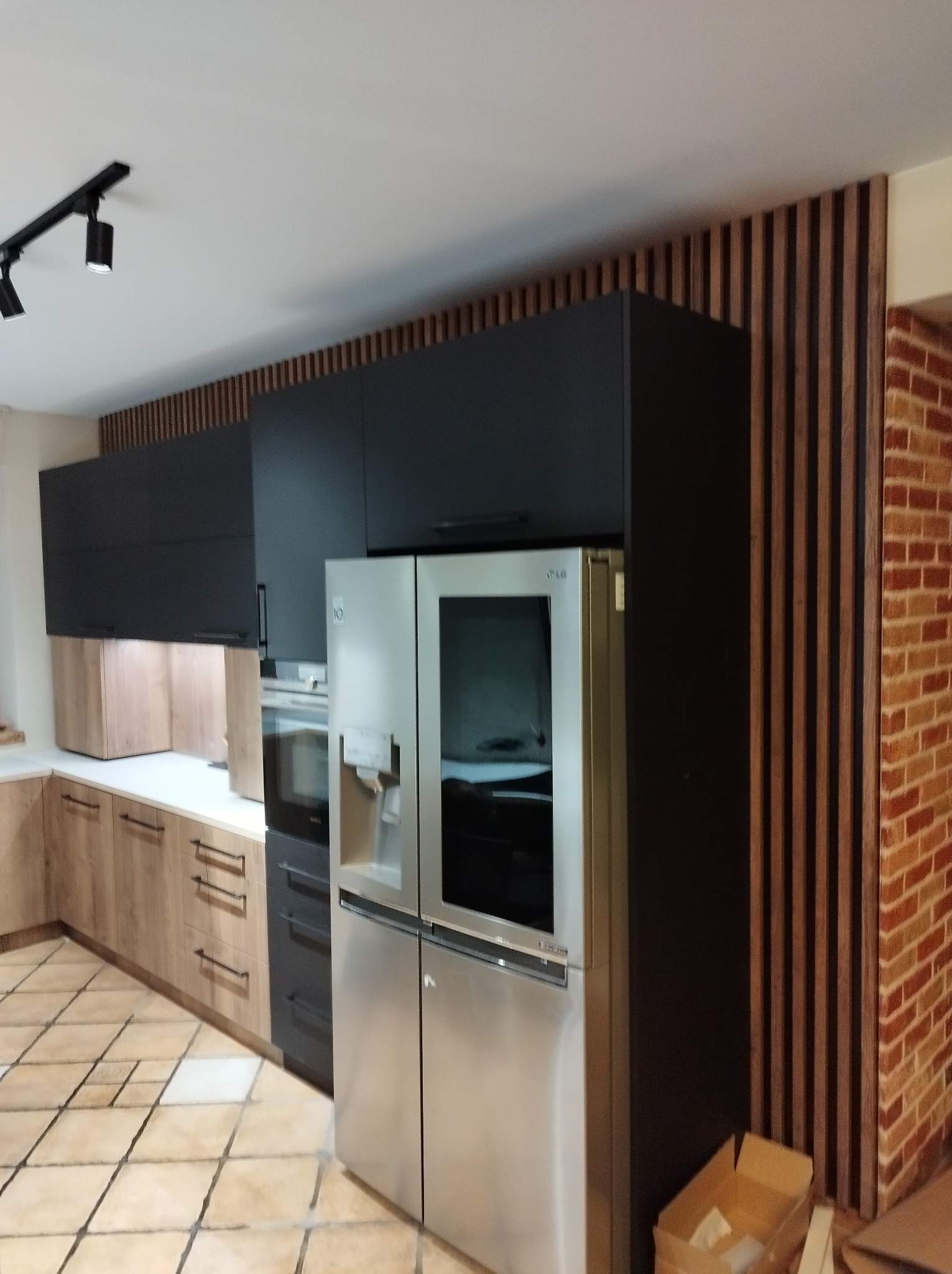 Nowoczesna kuchnia z czarnymi szafkami wiszącymi, drewnianymi lamelami na ścianie i dużą lodówką z przodu, oświetlona reflektorami sufitowymi.