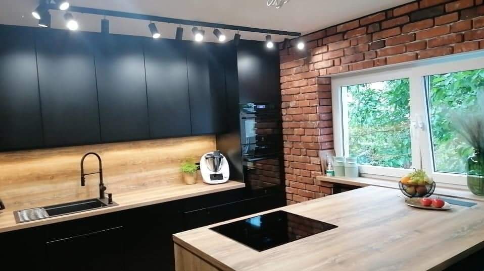 Nowoczesna kuchnia z czarnymi szafkami, drewnianym blatem, ścianą z czerwonej cegły i oświetleniem punktowym.