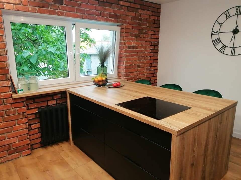 Wyspa kuchenna z drewnianym blatem i czarnymi frontami szuflad, płyta indukcyjna w blacie, ceglana ściana w tle, widok na zieleń za oknem.