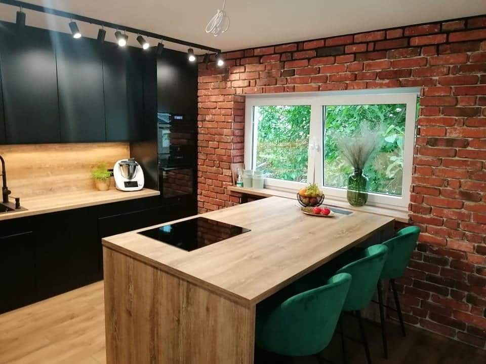 Nowoczesna kuchnia z czarnymi szafkami, drewnianym blatem, wyspą z płytą indukcyjną i ceglaną ścianą z oknem.