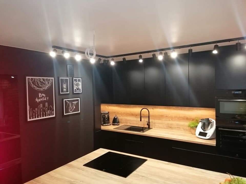 Nowoczesna kuchnia z czarnymi, matowymi szafkami, drewnianym blatem i oświetleniem punktowym; na ścianie grafiki w czarnych ramkach, na blacie ekspres do kawy i robot kuchenny.