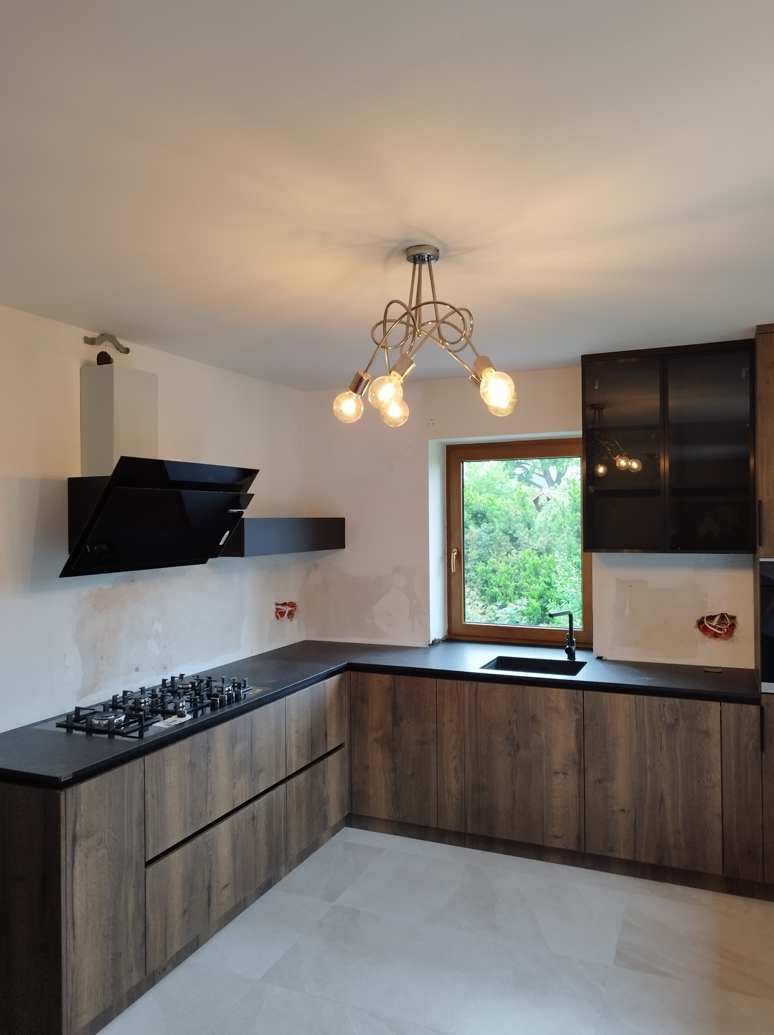 Kuchenna zabudowa w trakcie montażu: szafki dolne z frontami imitującymi drewno, czarny blat, płyta gazowa, okap i wiszące szafki z czarnymi, przeszklonymi frontami, widoczne instalacje elektryczne.