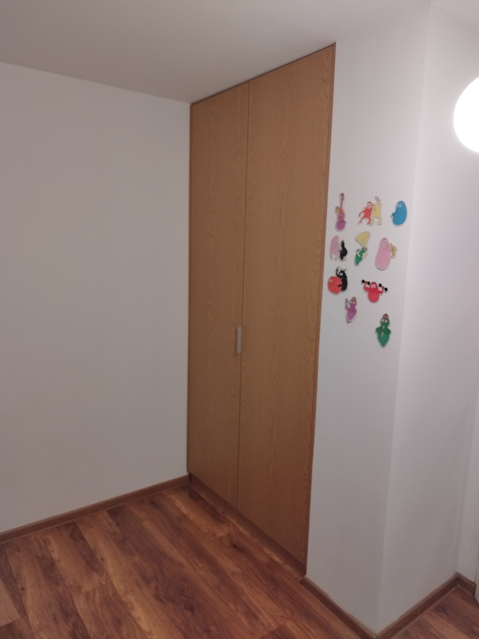 Wnętrze pokoju z drewnianą szafą wnękową, na której boku przyklejone są kolorowe naklejki z postaciami z bajek, podłoga wyłożona panelami w odcieniach brązu.