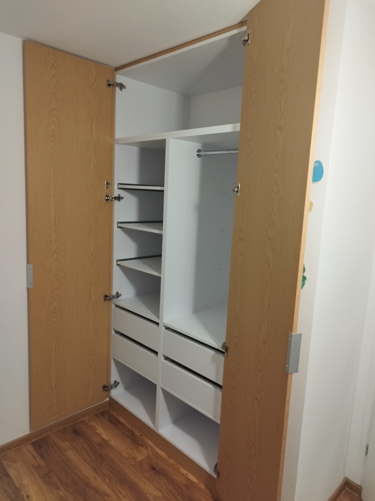 Otwarta szafa wnękowa z jasnego drewna i białymi półkami oraz szufladami, widoczne zawiasy i drewniana podłoga.