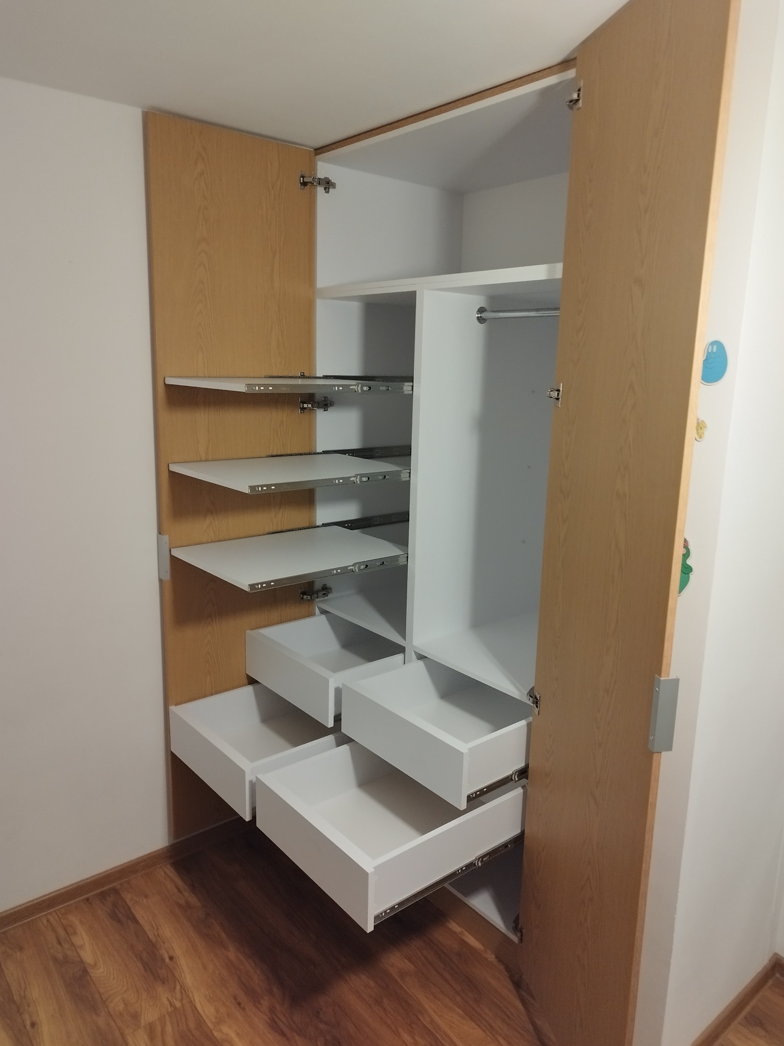 Otwarta szafa wnękowa z jasnego drewna z białymi półkami i szufladami na metalowych prowadnicach, widoczne zawiasy drzwiowe i drewniana podłoga.