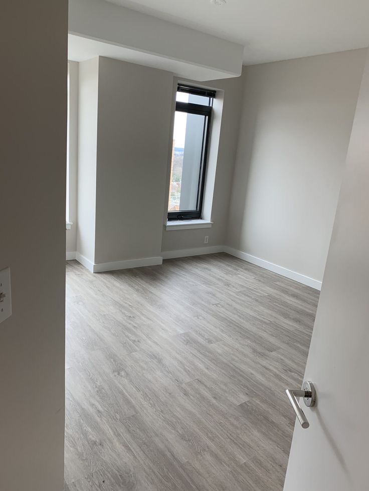 Pusty pokój z szarą podłogą imitującą drewno, świeżo pomalowanymi ścianami na jasny odcień szarości i czarną ramą okienną, widok zza uchylonych białych drzwi.