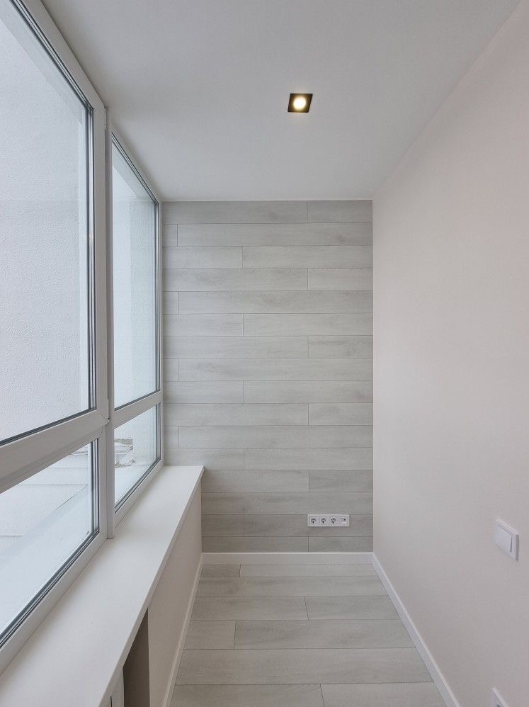 Wykończony balkon z szarą okładziną imitującą drewno na ścianie, podłodze i suficie, białe okno z widokiem na zewnątrz, oraz gniazdka elektryczne na dole ściany.