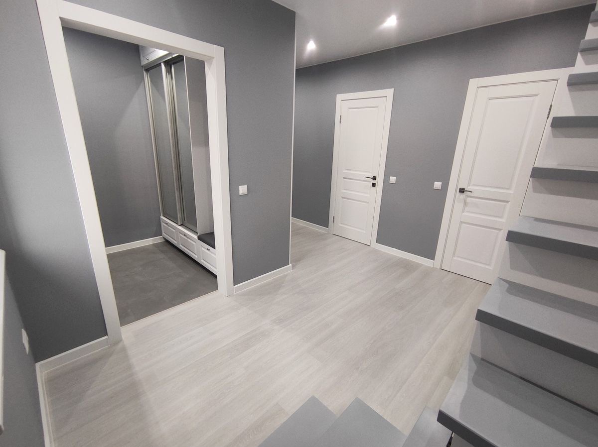 Szare wnętrze mieszkania z białymi drzwiami, podłogą imitującą drewno i zabudowaną szafą z siedziskiem w przedpokoju.