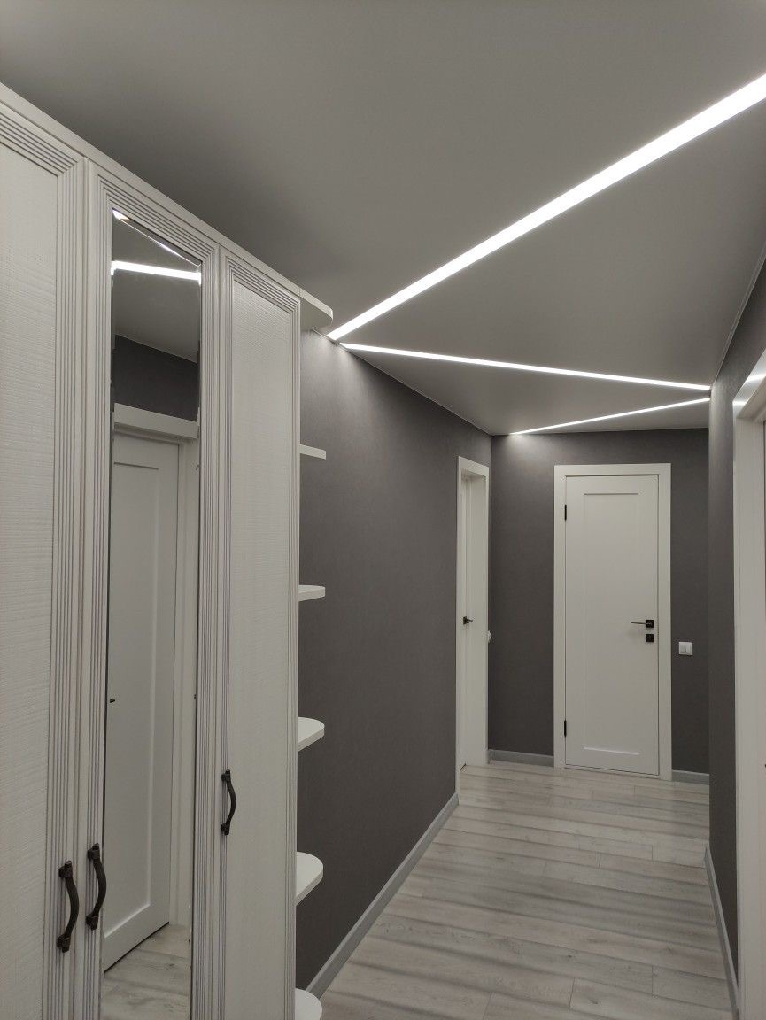 Korytarz z szarą ścianą, białymi drzwiami i szafą z lustrem, oświetlony liniowymi lampami LED na suficie.