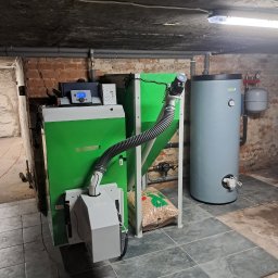 Idea Trader - Zainstalowany, zielony piec na pellet w piwnicy, z widocznym zasobnikiem pelletu, bojlerem i instalacją hydrauliczną na tle ściany z czerwonej cegły.