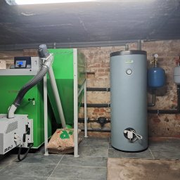 Idea Trader - Kotłownia z kotłem na pellet, zasobnikiem ciepłej wody i zbiornikami wyrównawczymi, widoczne rury instalacji grzewczej przy ceglanej ścianie.