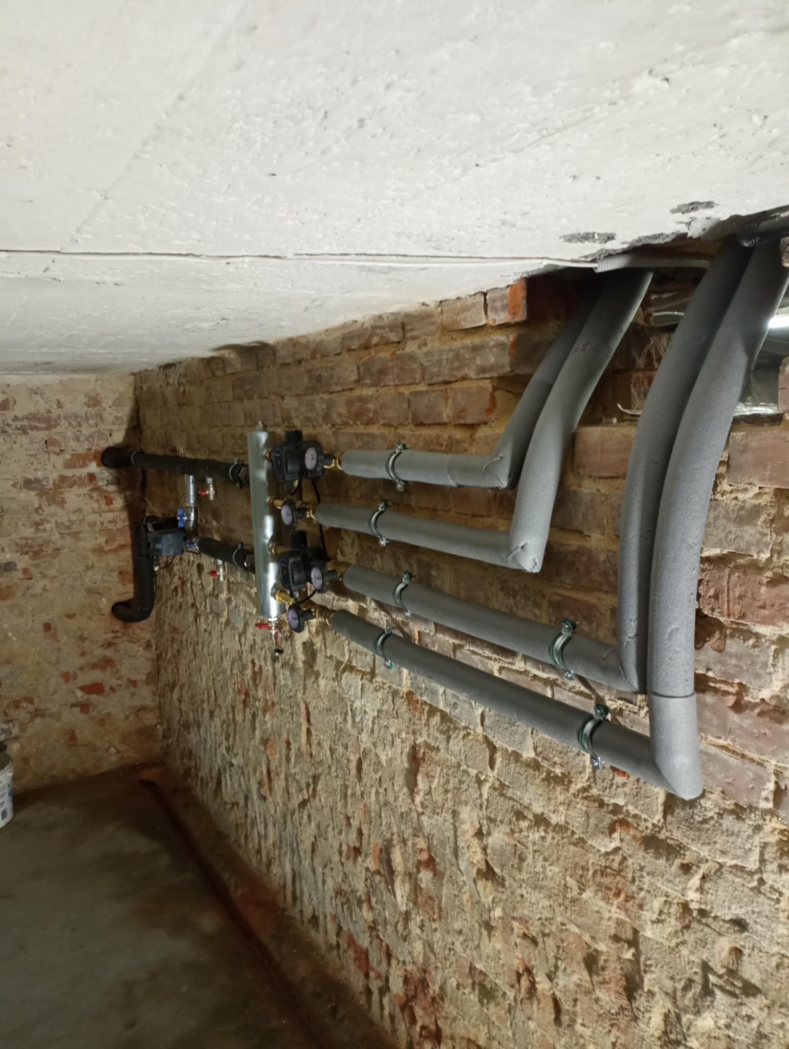 Instalacja centralnego ogrzewania z izolowanymi rurami na tle ceglanej ściany i betonowego sufitu, widoczne zawory i pompy obiegowe.