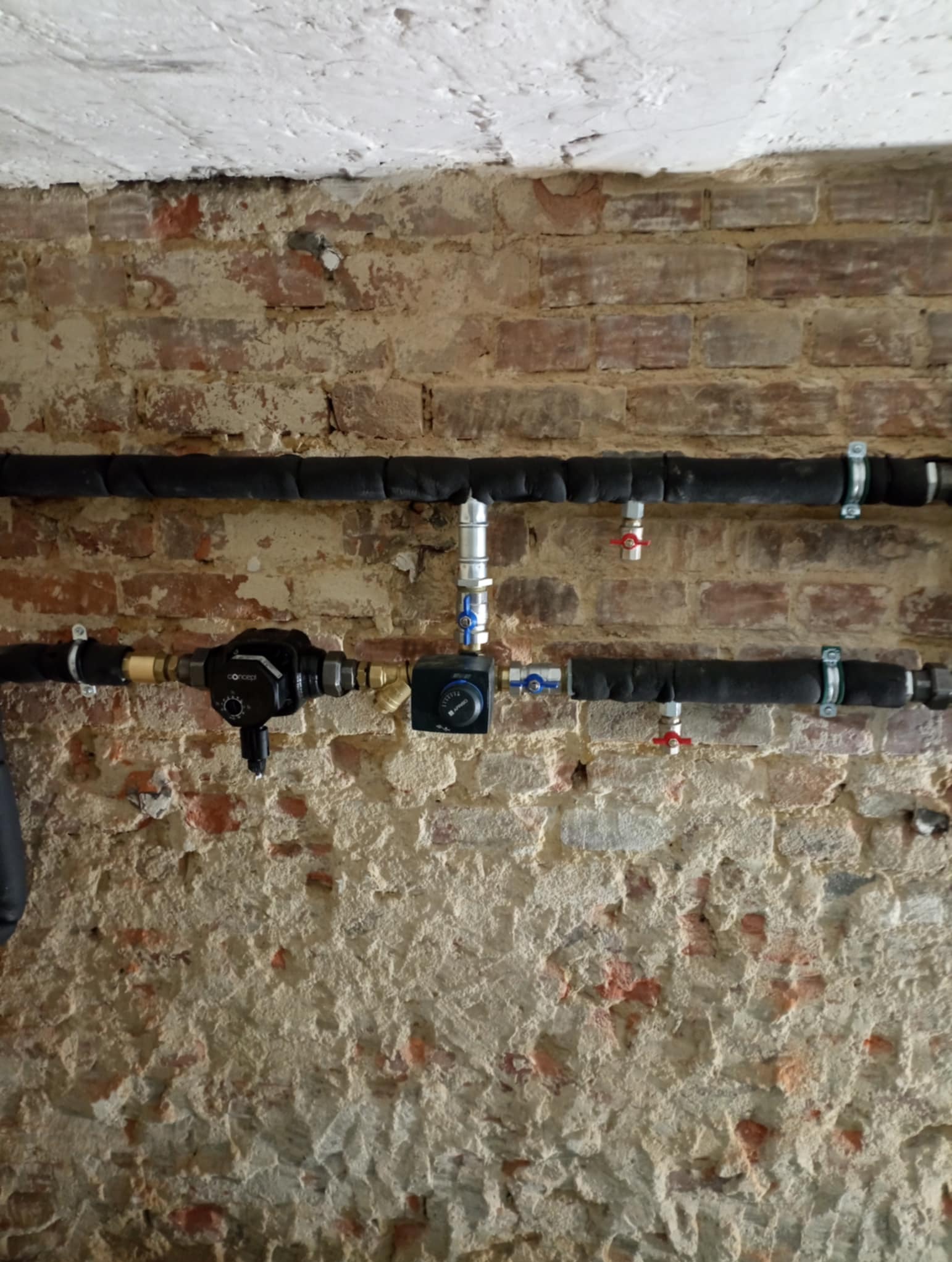 Fragment instalacji centralnego ogrzewania z zaworami i rurami w izolacji, zamontowany na starej, nierównej ścianie z cegły i zaprawy.