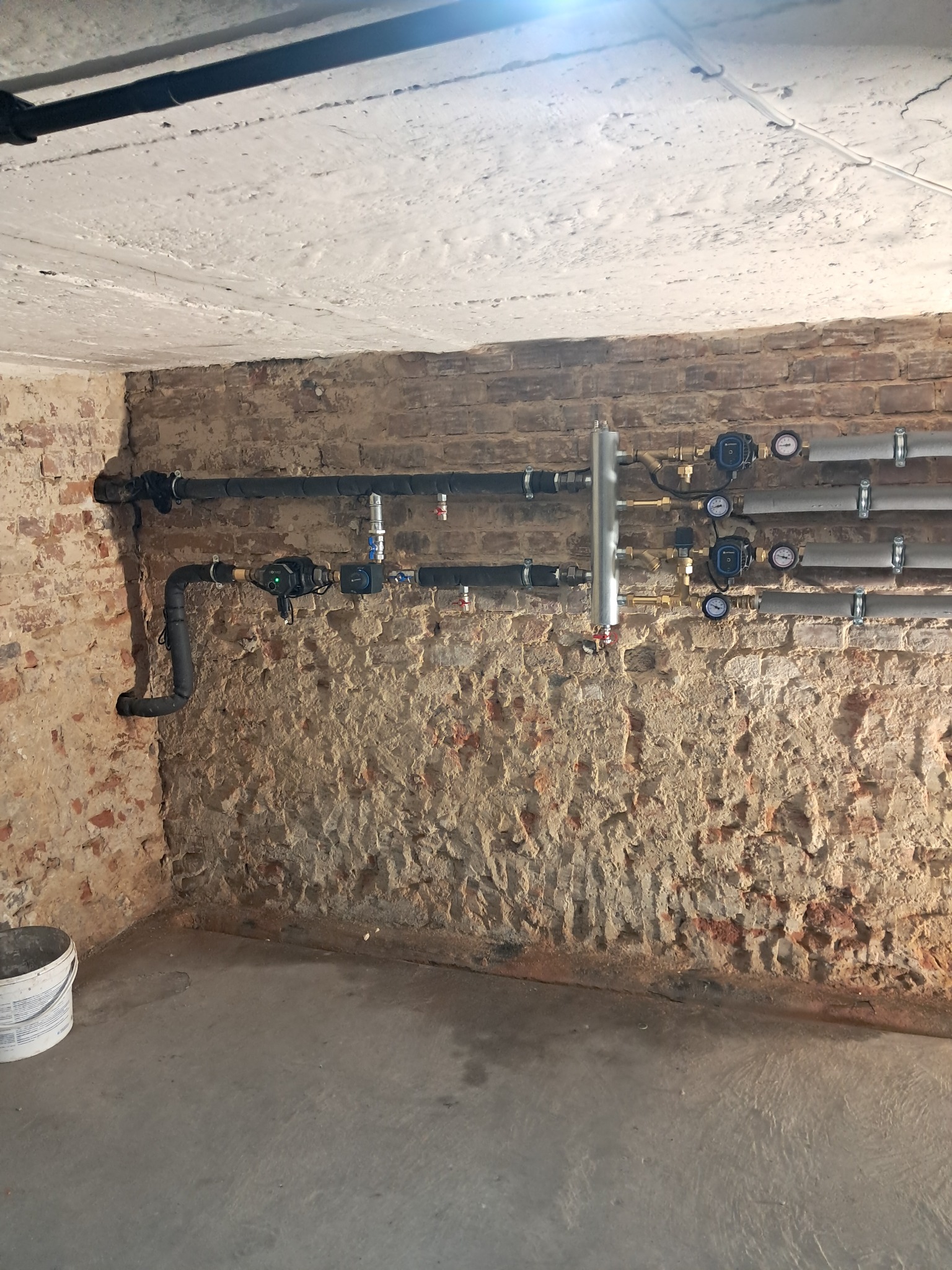 Instalacja rur i zaworów z manometrami na surowej, ceglanej ścianie w piwnicy, widoczny fragment podłogi i sufitu.