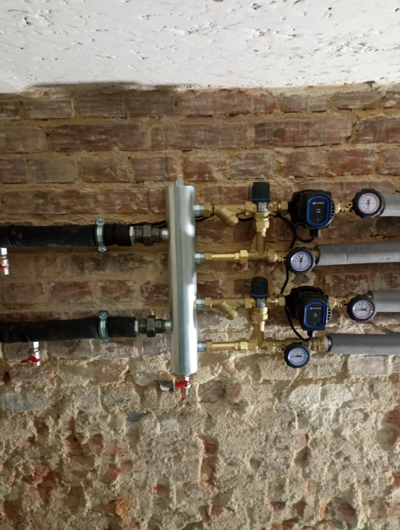 Instalacja hydrauliczna z rurami, zaworami, manometrami i pompami obiegowymi zamontowana na ścianie z surowej cegły.