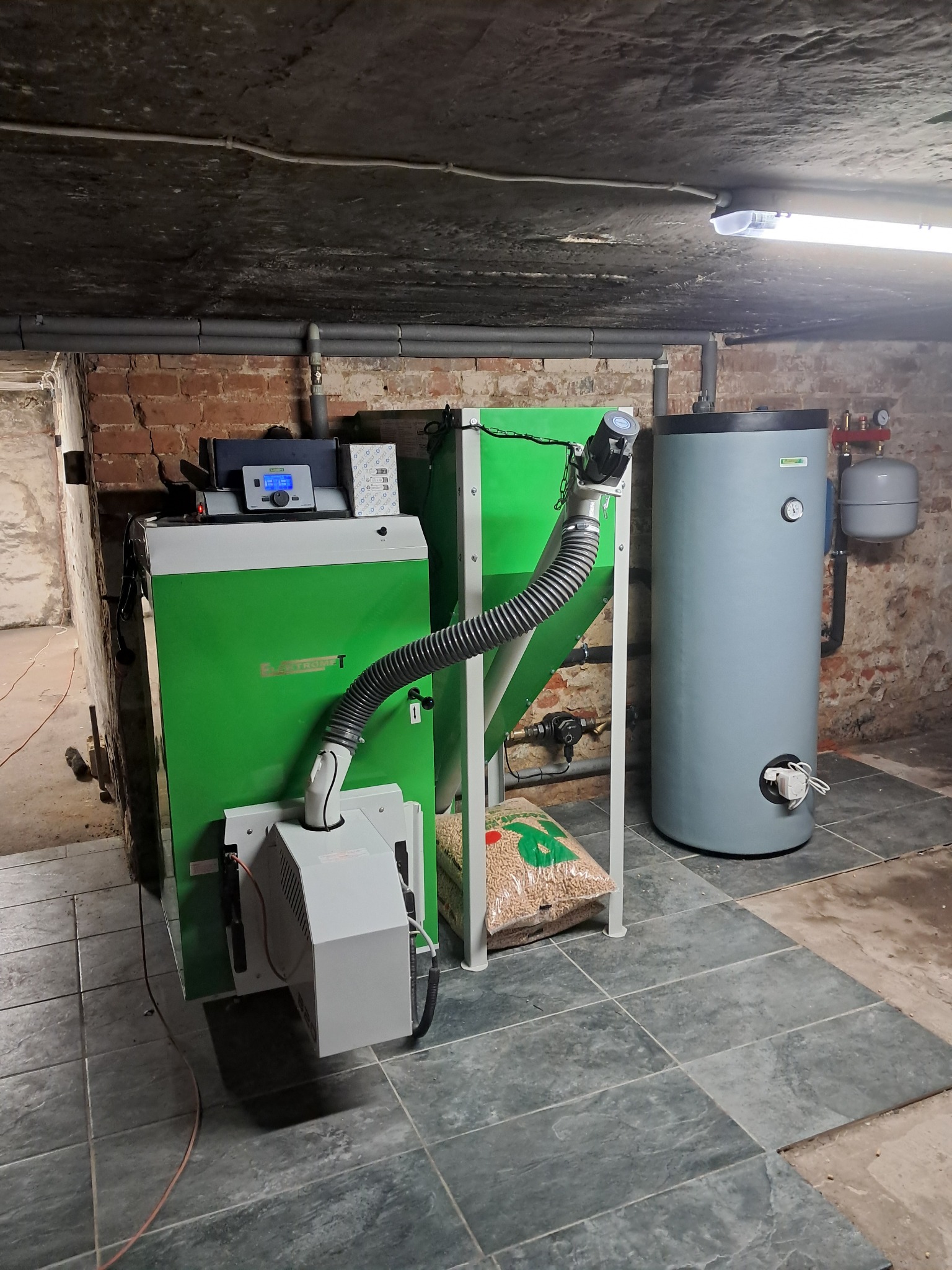 Zainstalowany, zielony piec na pellet w piwnicy, z widocznym zasobnikiem pelletu, bojlerem i instalacją hydrauliczną na tle ściany z czerwonej cegły.