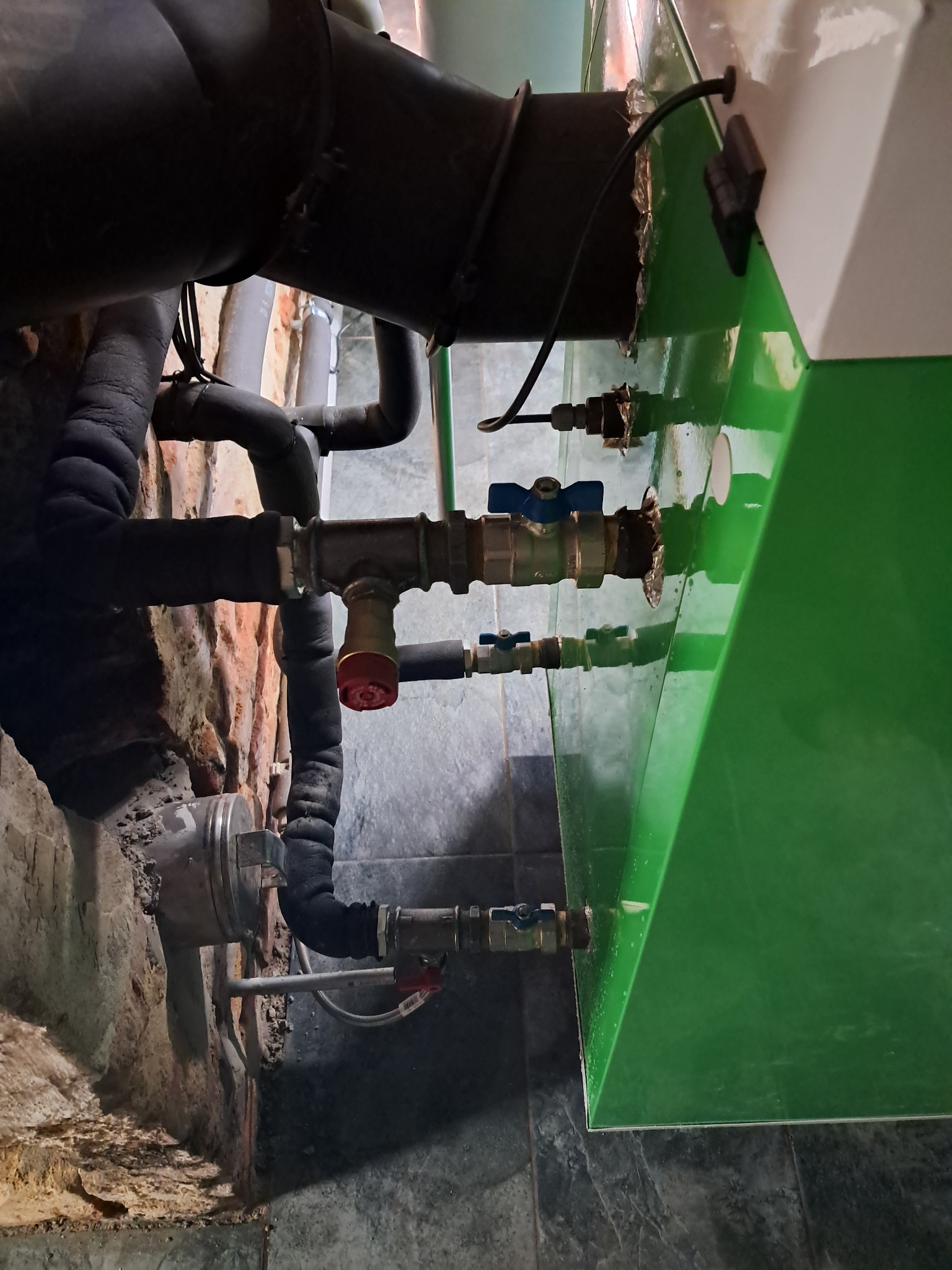 Fragment instalacji hydraulicznej z zaworami, rurami w izolacji i elementami kotła grzewczego przy ceglanej ścianie, widoczne ślady korozji i zużycia.