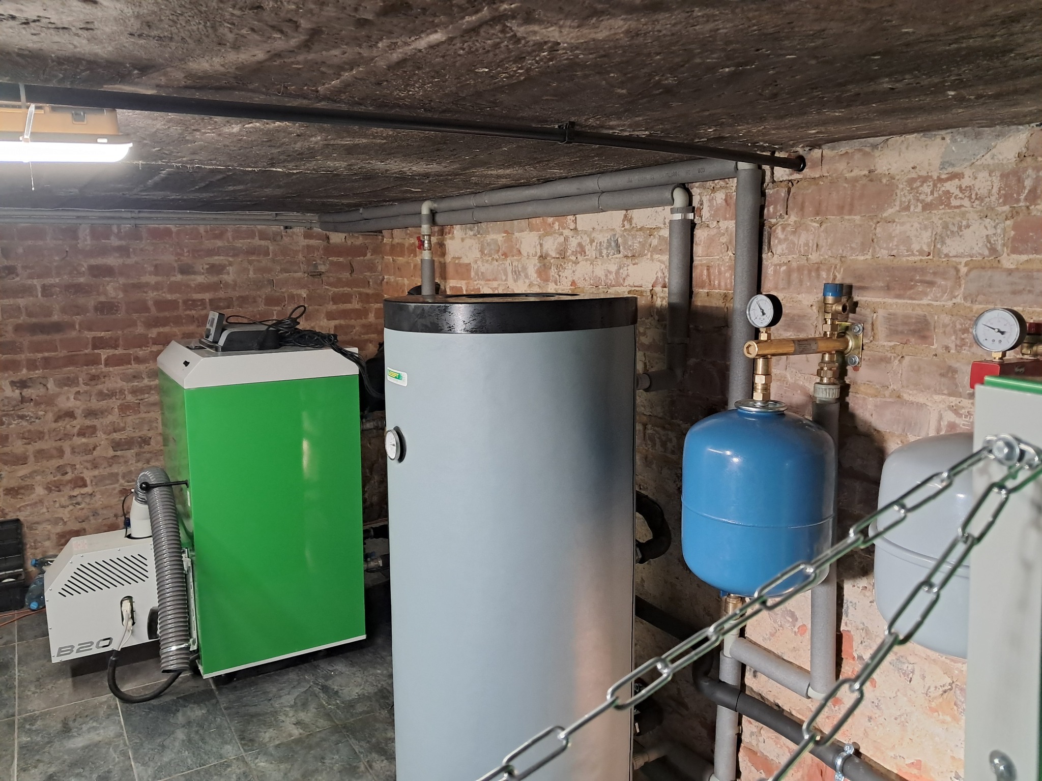 Kotłownia z piecem na pellet, zbiornikiem buforowym i instalacją hydrauliczną w piwnicy z ceglanym murem i betonowym sufitem.