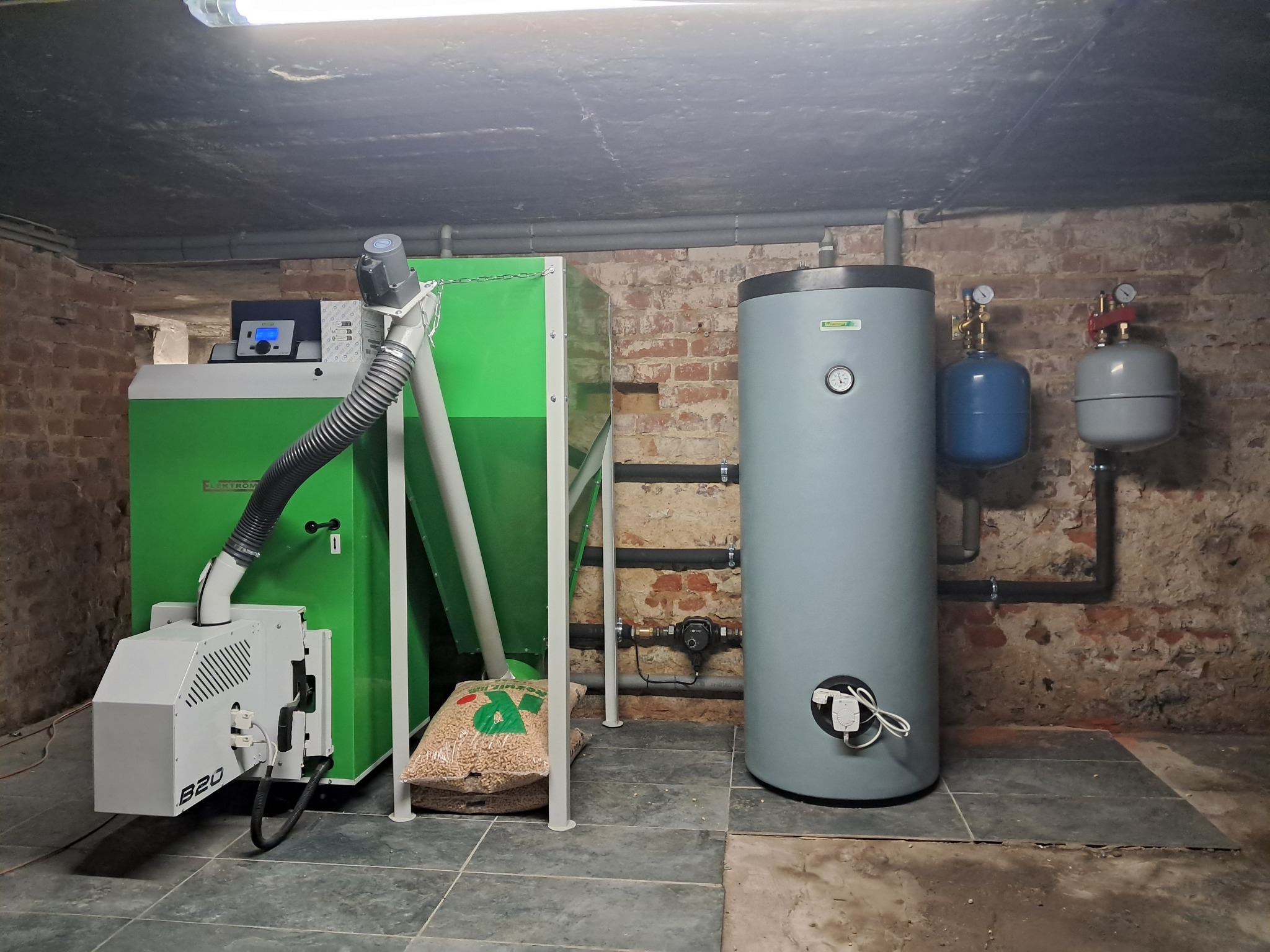 Kotłownia z kotłem na pellet, zasobnikiem ciepłej wody i zbiornikami wyrównawczymi, widoczne rury instalacji grzewczej przy ceglanej ścianie.