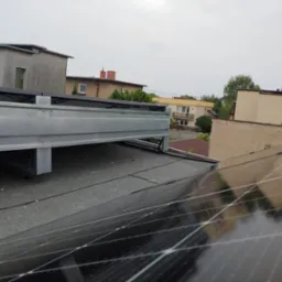 Montaż paneli fotowoltaicznych na płaskim dachu. Widoczna metalowa konstrukcja wsporcza i fragmenty modułów. W tle zabudowa miejska w pochmurny dzień.