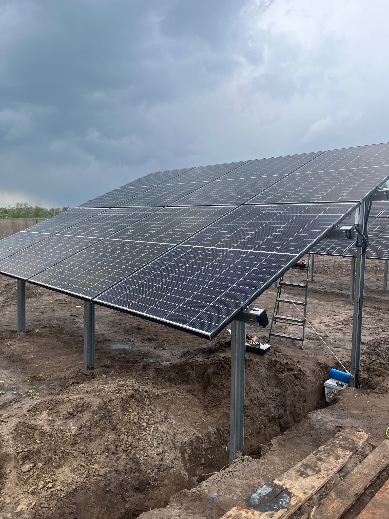 Grzęda paneli słonecznych na metalowych podporach, częściowo zamontowana na gruncie, widoczne wykopy ziemne i drabina, pochmurne niebo w tle.