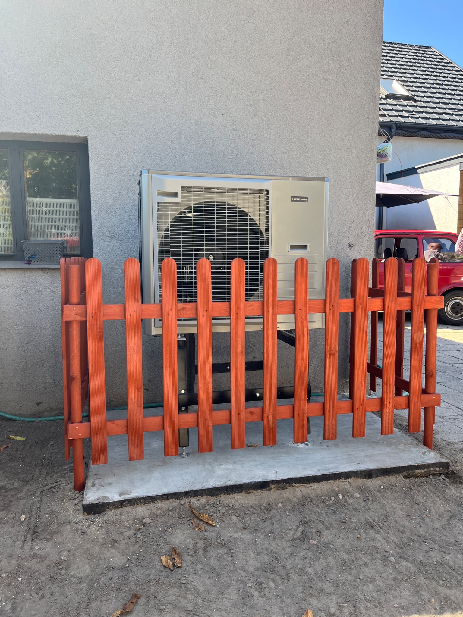 Zewnętrzna jednostka pompy ciepła Nibe AMS10 zabezpieczona niskim, pomarańczowym drewnianym płotkiem, zamontowana przy szarej ścianie budynku z oknem, w tle czerwony samochód