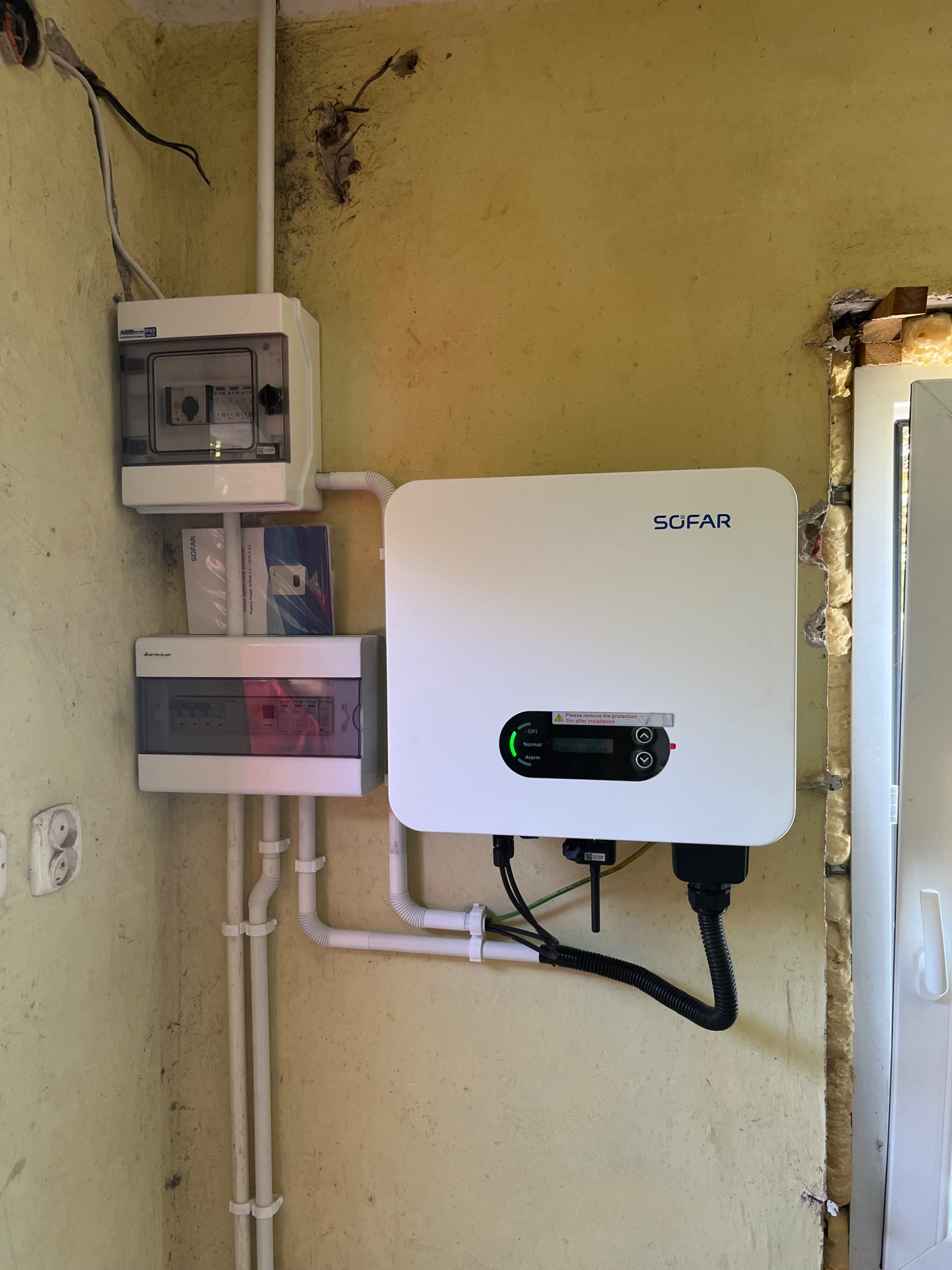 Zamontowany inwerter SOFAR z widocznymi przewodami, obok dwie skrzynki elektryczne na pomalowanej na żółto ścianie, widoczne uszczelnienie pianką montażową obok okna.
