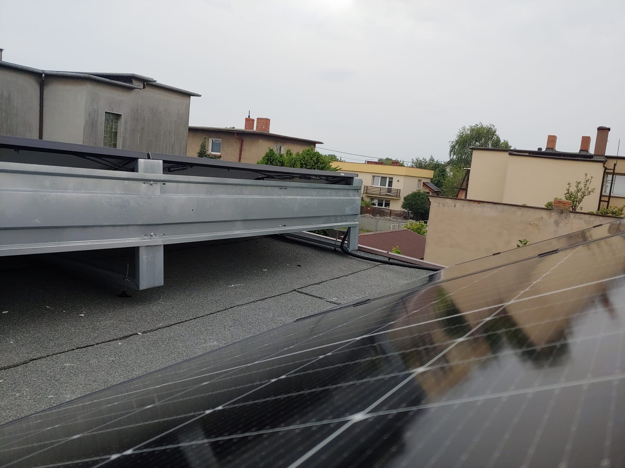 Montaż paneli fotowoltaicznych na płaskim dachu. Widoczna metalowa konstrukcja wsporcza i fragmenty modułów. W tle zabudowa miejska w pochmurny dzień.