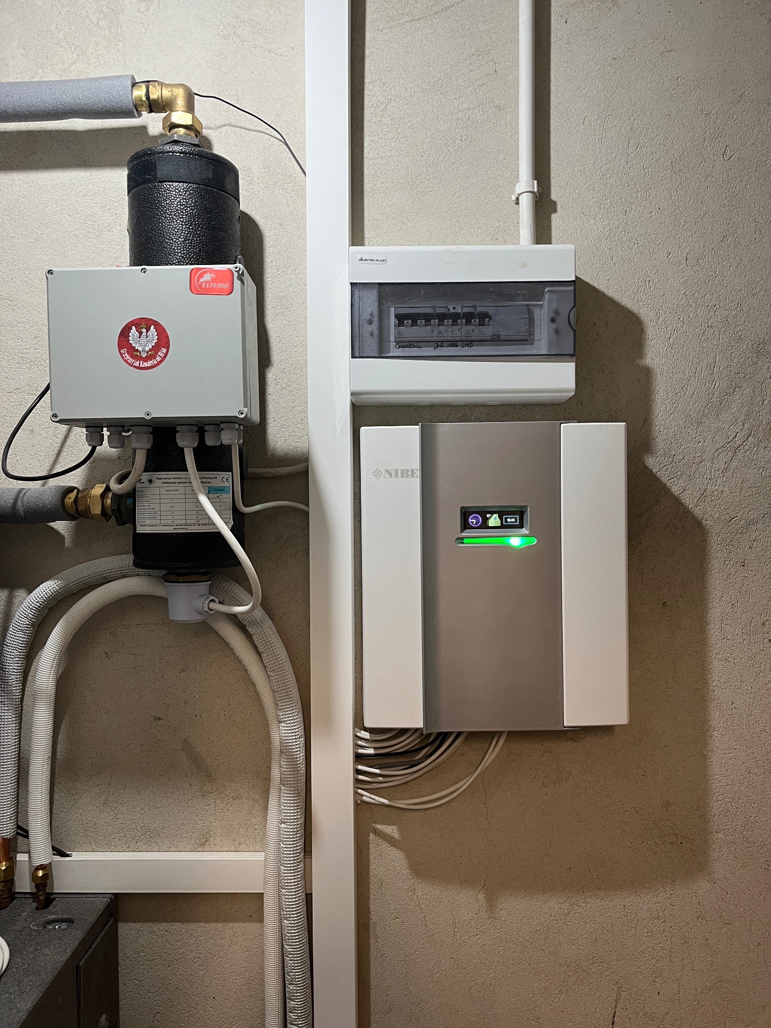 Szary moduł sterujący NIBE z zielonym wskaźnikiem LED, zamontowany na ścianie obok skrzynki elektrycznej i elementu instalacji grzewczej z rurami w izolacji. Widoczny fragment instalacji z czarnym...